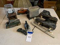 Full lot view showing all items on a table including gumball dispenser, two vintage metal toy cars, antique double door metal toaster, black clothes iron, leather and metal ice skates with blade covers.