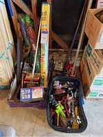 Photo showing a box and a basket containing assorted hand tools, garden accessories, marbles, weed control, and kites.