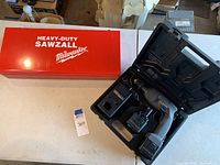 Closed red metal case labeled Heavy-Duty Sawzall Milwaukee and open black plastic case with cordless drill inside