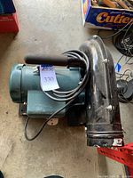 Side view showing the portable blower unit, featuring a curved black exhaust with protective mesh grid and green motor housing with foam handle grip and attached power cable.