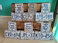 Top view showing all 6 pairs of white and blue 1973 Ontario license plates arranged on wooden supports and a teal floor; various plate numbers visible.