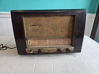 Front view of antique Philips radio showing dark wooden casing, worn fabric speaker grille, tuning dial with station markings, and three control knobs.