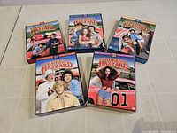 Five boxed DVD sets of Dukes of Hazzard seasons 1 to 5 arranged on a flat surface, showing front cover artwork with characters and General Lee car.