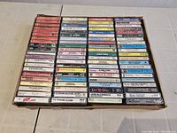 Top-down photo showing the box containing approximately 80 cassette tapes stacked in rows, with visible labels and covers including soundtracks and artist names.