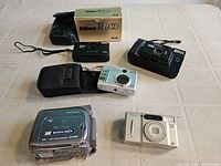 Overview of five cameras on table: Nikon AF600 box and pouch, Optex film camera, Creative DC-Cam 32002, Nikon Nuvis 300, Canon ZR500 camcorder