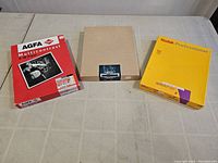 Top-down view of three unopened boxes of photo paper from AGFA, MCIV, and Kodak Professional on table.
