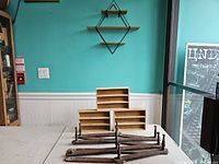 Photo showing metal accordion wall shelf mounted on turquoise wall with two wooden shelves. Three wooden compartment boxes and metal accordion wall shelf frame pieces on table in front.