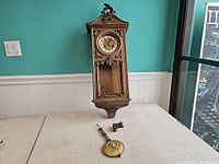 Full view of wall clock case standing on table with pendulum and decorative wooden piece in front of it.