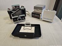 Photo showing the Polaroid Automatic 250 Land Camera, two boxed kits labeled Polaroid Close-Up Kit (9513A) and Polaroid Portrait Kit (9514A), and a black plastic tray with metal frame on a table.