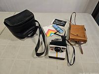 Full lot overview showing Polaroid OneStep camera in center with two leather carrying cases (black and brown), manuals and film pack on table