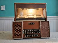 Front and side view of the 1960s tabletop diner jukebox showing illuminated top display, letter and number selector buttons, speaker grille, and textured brown casing with wear and scratches.
