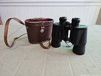 Photo showing Tasco 10 x 50 binoculars standing upright next to brown leather carrying case with strap on a light-colored table.