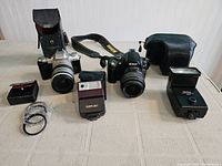 Front view of lot showing Pentax MZ50 camera with attached lens and leather case, Nikon D40 with Nikkor lens and strap, Canon Speedlite 244T, Vivitar flash and additional accessories spread on table