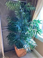 Full view of the artificial bush with long green palm-like leaf stems in wicker basket planter next to window.