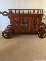 Front view of cherry stagecoach tea cart with glass-paneled cabinet, spindle railings, decorative carving and wood wheels