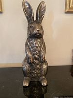 Front view of decorative metal rabbit statue on black surface with wall and framed art in background