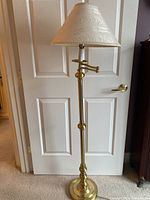 Full front view of brass floor lamp with fabric shade in front of white paneled door, showing lamp extended upwards and straight.