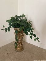 Gold-colored decorative floor plant stand with three cherubs holding up a round bowl shown with a green leafy plant inside.