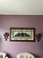 Wall view showing the ornate framed art hung centrally between two decorative matching wall shelves on a purple-painted wall above couch backs.