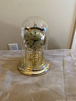 Front view of the gold-colored clock under a clear glass dome, showing the circular dial with Roman numerals and the pendulum balls at the base.