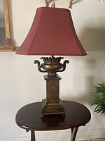 Ornate electric table lamp with red fabric shade and bronze/gold-tone decorative base featured frontally on a round wood table.