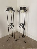 Pair of black metal standing candleholders with scrollwork and clear glass tops containing battery operated candles placed on carpeted floor against a white wall.