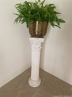 Full view of white plaster column with decorative fluted shaft and Corinthian style capital topped with a brass colored planter holding a fern plant.