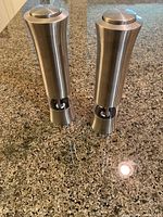 Two silver-colored battery operated grinders standing upright on a granite countertop showing the front and side profile.