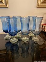 Full set of six blue and clear stemmed glasses arranged in a row on a reflective black surface with framed art in the background.