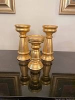 Front view of three gold colored candleholders, showing two taller ones and one shorter in front, reflecting on glossy surface.