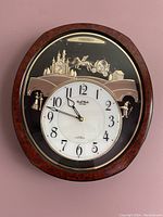 Full view of the oval musical wall clock with wood frame, white face, and decorative upper relief depicting castle and horse carriage.