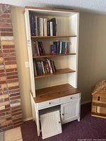Full view of the laminated wooden bookcase showing three upper shelves filled with books, two drawers below, and a bottom cabinet with one detached door lying on the floor.