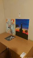 Three canvas art pieces on a wooden surface against a beige wall, includes a floral painting, lighthouse sunset, and a smaller dark-themed painting.