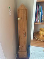Tall, narrow wooden plywood CD storage cabinet shown closed and positioned by a wall and furniture with a triangular top and bottom drawer.