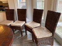 Four dark brown woven wicker chairs with beige seat cushions next to a wooden dining table in a kitchen area. Light from windows behind the chairs. Cushions appear used and need cleaning.