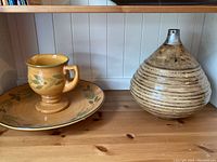 Two pottery pieces on a wooden shelf: a ceramic cup and plate with leaf design, and a large bulbous ceramic vase.