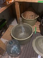 View of two winemaking demijohns in protective baskets and a standalone 21-inch glass bottle on basement floor.