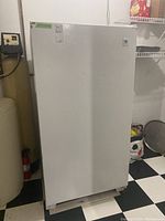 Side and front view of white upright freezer in basement with checkered floor, showing closed door with some hardware and labels.