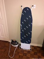 Full view of ironing board standing up against a wall, showing blue fabric cover with multiple 'DeLonghi' logos, metal legs, and iron rest at bottom.