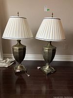 Pair of urn-shaped resin table lamps with pleated cream fabric shades, decorative finials, placed on dark wood floor against neutral wall.