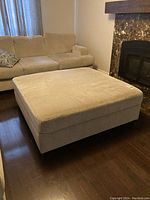 Front and perspective view of a beige upholstered ottoman placed in a living room with a sofa and dark wooden floor.