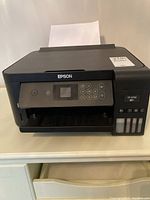 Front view showing the Epson logo, control panel, output tray, and paper loaded in the top tray.
