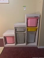 Front view of painted wooden cabinet with pink, yellow, and gray plastic storage bins inserted in stepped compartmentalized shelving.