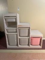 Front and side angle view of a white painted wood step-style storage cabinet with multiple plastic bins (white and one pink). The cabinet has compartments for the bins and measures approximately 37 x 17 x 36 inches.