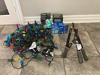 One Fiskars pair of garden trimmers with foam grip handles next to multiple sets of Christmas lights and their boxes on tile floor.