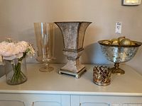 Wide shot of three vases and a large gold pedestal bowl filled with Christmas ball ornaments, plus a silver candle holder on a white table surface.
