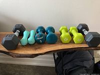 All four pairs of hand weights shown together on a wooden surface, arranged left to right: black 8 lb hexagonal weights, light blue small vinyl weights, teal LifeGear 5 lb weights, lime green 5 lb weights.