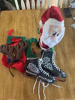 Plush Santa, ice skate ornaments, elf hat, and reindeer headband on a wood table.