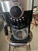 Front and top view of the black Cuisinart coffee maker with glass carafe on a wooden stool, showing control dial and buttons