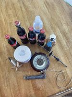Mixed items including six Coca-Cola glass bottles, a roll of white yarn or twine, a round barometer, a carved wooden object, a metal folding tool or knife, a metal rod with handle, small plastic bottle, and some string on a wooden table.